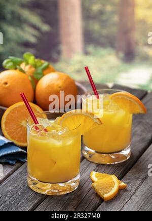 Orangensaftcocktail mit Orangenscheiben und frischen Orangen auf einem alten Holztisch mit Naturkulisse Stockfoto