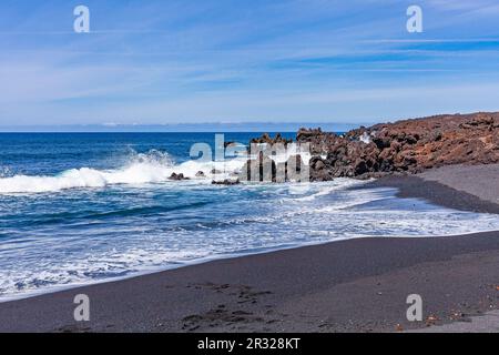 Leuchtender Kontrast aus weißem Meerschaum der Wellen, die mit Sand und schwarzer verfestigter Lava an die Küste Rollen. Stockfoto