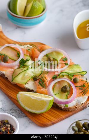 Offene Sandwiches mit gesalzenem Lachs, Frischkäse, Zwiebeln, Kapern, Gurke und Mikrogreens auf Holzbrett mit weißem Küchentisch. Stockfoto