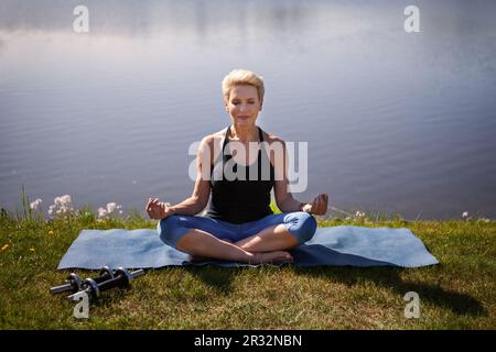 Friedliche reife Frau übt Yoga im Freien. Sportlerin in Lotusposition vor dem Hintergrund des Naturwassers. Aktiver Lebensstil und Heilung Stockfoto