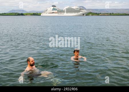 Bantry, West Cork, Irland. 22. Mai 2023. Der erste luxuriöse Kreuzfahrtschiff des Jahres, „Seabourn Ovation“, kam heute Morgen in Bantry Inner Harbour an, mit 600 Passagieren. Sie segelt heute Abend um 5pm Uhr. Paudie Holden und Gavin O'Shea aus Bantry nehmen ihr morgendliches Bad mit dem Schiff im Hintergrund. Kredit: AG News/Alamy Live News Stockfoto