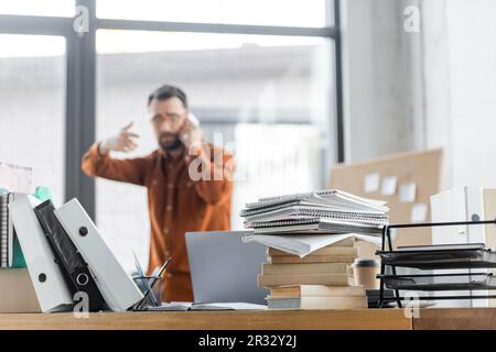 Selektiver Fokus auf Büroschreibtische mit einem Stapel von Büchern, Notizbüchern, Ordnern und Laptops, die Geschäftsleuten beim Telefonieren über Mobiltelefon und sol nahe stehen Stockfoto