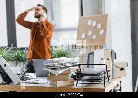 Geschäftiger und müder Geschäftsmann, der sich mit einem Smartphone unterhält und das Problem in der Nähe von Korkplatten mit Haftnotizen und einem Schreibtisch mit Notizbüchern, Büchern und Pappe löst Stockfoto