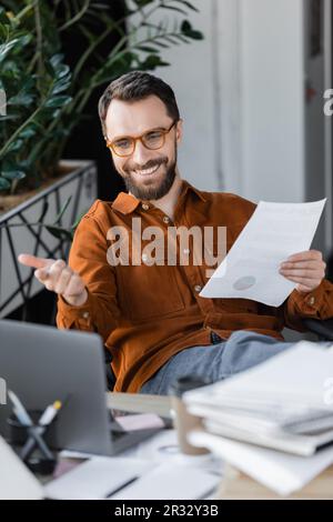 Freudiger Manager mit Bart in stilvoller Brille und Hemd, der ein Dokument in der Hand hält und während einer Videokonferenz auf den Laptop zeigt, in der Nähe verschwommener Dokumente auf der Arbeit Stockfoto
