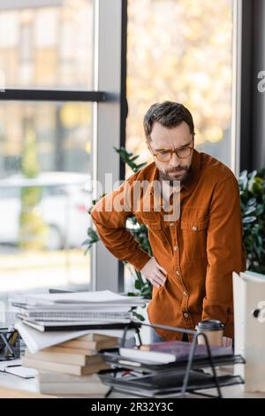 Ein bärtiger Geschäftsmann in einer Brille, der mit der Hand auf der Hüfte steht und sich neben dem Pappbecher auf dem Schreibtisch viele Bücher, Notizbücher und Ordner ansieht Stockfoto