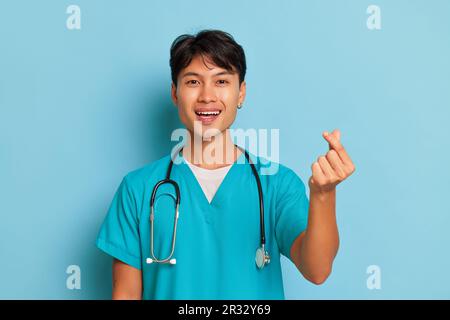 Netter junger Arzt steht auf blauem Hintergrund, die Finger seiner linken Hand in Herzgesten falten, vor der Kamera lächeln, professionelles Menschenkonzept, Kopie Stockfoto
