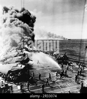 Die USA Navy Light Aircraft Carrier USS Belleau Wood (CVL-24) brennt achtern, nachdem sie von einem Kamikaze getroffen wurde, während sie am 30. Oktober 1944 in Luzon (Philippinen) operierte. Die Besatzung des Cockpits bewegt unbeschädigte Grumman TBM Avenger-Flugzeuge des Torpedo-Geschwaders 21 (VT-21) von den Flammen weg, während andere die Brände bekämpfen. Die USS Franklin (CV-13), die ebenfalls während des Kamikaze-Angriffs getroffen wurde, ist in der Ferne in Flammen. Stockfoto