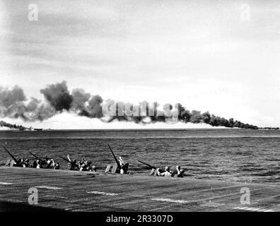 Mitglieder der Marine-Einheit an Bord der USA Der Navy-Flugzeugträger USS Enterprise (CV-6) schützt ihre Augen vor der Sonne, während sie den Himmel nach Kamikaze-Flugzeugen scannen, die aus der Sonne springen. Beachten Sie, dass die Fluggesellschaften USS Franklin (CV-13), Left und USS Belleau Wood (CVL-24) nach dem Zusammenstoß mit Kamikaze-Flugzeugen brennen. Stockfoto