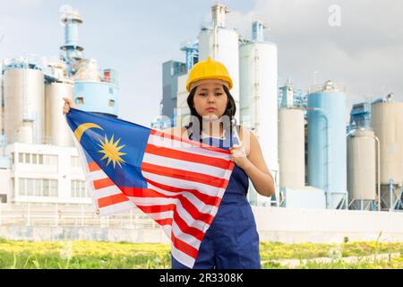 Traurige junge Frau im Helm mit der Flagge Malaysias vor dem Hintergrund der Fabrik Stockfoto