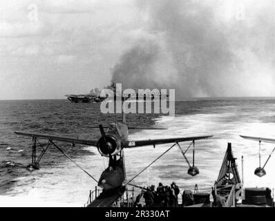 USS Ticonderoga (CV-14) brennt, nachdem sie am 21. Januar 1945 von einem „Kamikaze“-Angriff vor Formosa getroffen wurde. Fotografiert von der USS Miami (CL-89). Ein Vought OS2U "Kingfisher" Wasserflugzeug befindet sich auf dem Steuerbordkatapult des Kreuzers im Vordergrund. Als Japan Ende 1944 mit einer Niederlage konfrontiert war, entschied es sich, US-Schiffe mit Selbstmordattentaten, bekannt als Kamikaze, zu zerstören. Diese Angriffe waren eine starke physische und psychologische Waffe und versenkten insgesamt 47 Schiffe zu einem Preis von mehr als 3000 Piloten und Flugzeugen. Ende 1944 war die US-Marine so groß, dass die Verluste unbedeutend waren, und sie änderte den Kurs von t nicht Stockfoto