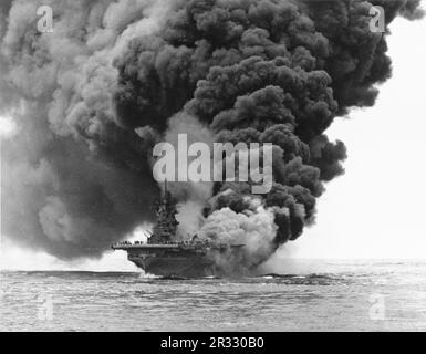 Die USA Navy Aircraft Carrier USS Bunker Hill (CV-17) brennt nach dem Zusammenstoß von zwei „Kamikaze“-Selbstmordflugzeugen vor Okinawa am 11. Mai 1945. Fotografiert von USS Bataan (CVL-29). Der Streik fing voll betankte und bewaffnete Flugzeuge an Deck und löste sehr gefährliche Brände aus. Obwohl das Schiff schwer beschädigt war, ging es nicht unter und kehrte zur Reparatur in die USA zurück. Das Schiff wurde 1966 stillgelegt und 1973 verschrottet. Stockfoto