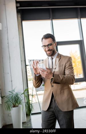Ein freudiger Manager mit Bart in stilvoller Business-Kleidung und Brille winkte während eines Videoanrufs auf dem Handy in der Nähe des Flipcharts und einer Topfpflanze in moderner Form Stockfoto