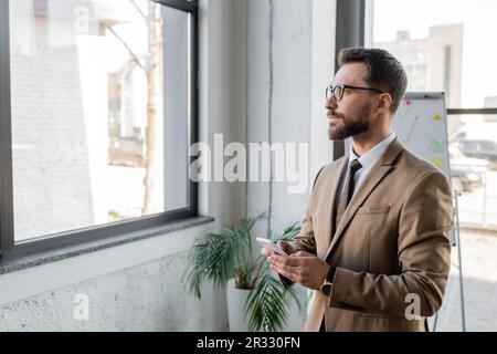 Ein seriöser, bärtiger Geschäftsmann in beigefarbenem Blazer und einer Brille, der sein Smartphone hält, während er durch das Fenster blickt und in die Nähe des Flipcharts und der vergossenen pla denkt Stockfoto