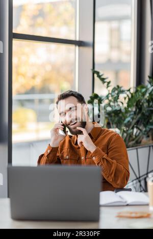 Lächelnder Geschäftsmann, der Bart berührt und mit dem Handy in der Nähe des Notebooks spricht und ein verschwommenes Notebook neben dekorativen Pflanzen im Hintergrund im zeitgenössischen Stil hat Stockfoto