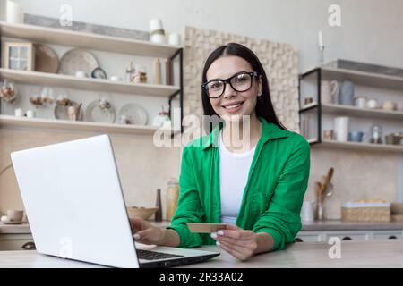 Eine junge Freiberuflerin arbeitet zu Hause in der Küche an einem Laptop und benutzt eine Kreditkarte. Er sieht mit einem Lächeln in die Kamera. Stockfoto
