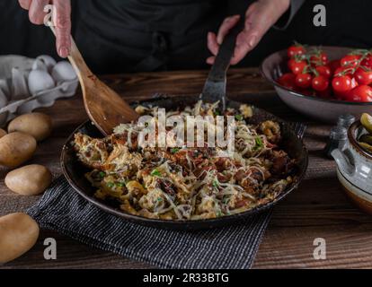 Bauernfrühstück mit Bratkartoffeln, Speck, Eiern, Schnittlauch und Käse. Serviert in einer rustikalen Eisenpfanne auf einem Holztisch. Stockfoto