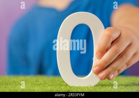 Nummer 0 in der Hand. Hand mit weißer Nummer Null über grünem Gras und lila Hintergrund, Konzept von Null-Emission CO2, Zinssätze, Nullpunktzahl in Stockfoto