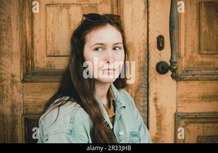 Eine schöne junge Frau, die eine Jeansjacke trägt und an einer Holztür steht. Zeitgenössische Mode und traditionelle Umgebung. Stockfoto
