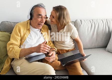 Älterer älterer Mann, der Musik über kabellose Kopfhörer hört, Frau hebt den Ohrhörer und spricht im Ohr des Ehemanns Stockfoto