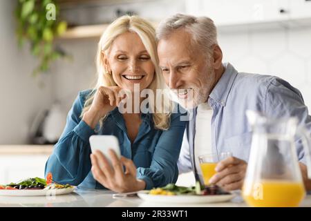 Glückliches Seniorenpaar, das während des Frühstücks in der Küche ein Smartphone benutzte Stockfoto