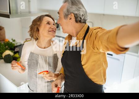 Genießen Sie das gemeinsame Kochen. Liebevolles Seniorenpaar, das Selfie mit Pfefferscheiben macht und sich die Küche anschaut Stockfoto