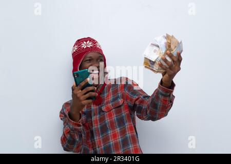 Aufgeregter junger afrikanischer Mann, der Bargeld in der Hand hat und sein Telefon zum Spaß benutzt Stockfoto