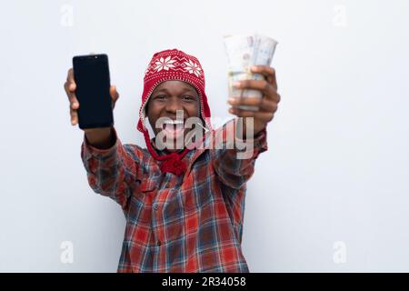 Aufgeregter junger afrikanischer Mann, der Bargeld in der Hand hat und sein Telefon zum Spaß benutzt Stockfoto