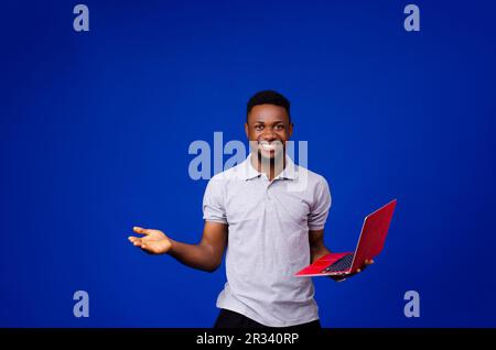 Ein junger afrikanischer Mann, der mit einem Laptop auf einem isolierten blauen Hintergrund steht und sich aufregt Stockfoto