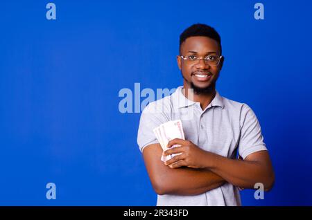 Ein junger, gutaussehender Afrikaner, der lächelt, während er Bargeld in den Armen hält und auf einem blauen, isolierten Hintergrund steht Stockfoto