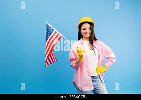 Ein Mädchen in einem Bauhelm und Handschuhen mit amerikanischer Flagge steht auf blauem Hintergrund Stockfoto