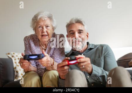 Ein erwachsener Mann spielt Videospiele mit seiner älteren Mutter und amüsiert sich. Stockfoto