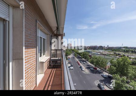 Lange Terrasse eines urbanen Wohnhauses mit Metallgeländer und durchsichtigem Glas mit Blick auf die Stadt und eine große, von grünen Bäumen gesäumte Straße Stockfoto