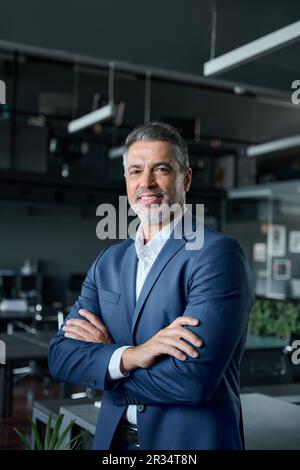Ein glücklicher Geschäftsmann mittleren Alters steht im Büro, mit gekreuzten Armen, vertikales Porträt. Stockfoto