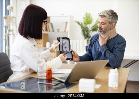 Ärztin mit Stethoskop und Patient in Nackenstütze sitzt am Schreibtisch mit Digital-Tablet in der Arztpraxis. Radiologe zeigt Zerv Stockfoto