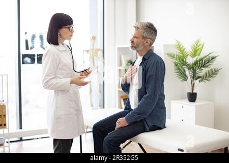Besorgter, grauhaariger Mann, der auf der Untersuchungscouch sitzt, mit der Hand auf der Brust, während eine aufmerksame Frau im Mantel des Arztes ein Stethoskop hält. Männlicher Patient, der dem Facharzt in der Sprechstunde Symptome beschreibt. Stockfoto