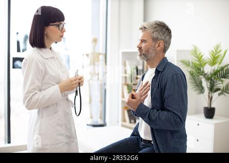 Besorgter, grauhaariger Mann, der auf der Untersuchungscouch sitzt, mit der Hand auf der Brust, während eine aufmerksame Frau im Mantel des Arztes ein Stethoskop hält. Männlicher Patient, der dem Facharzt in der Sprechstunde Symptome beschreibt. Stockfoto