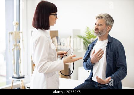 Besorgter, grauhaariger Mann, der auf der Untersuchungscouch sitzt, mit der Hand auf der Brust, während eine aufmerksame Frau im Mantel des Arztes ein Stethoskop hält. Männlicher Patient, der dem Facharzt in der Sprechstunde Symptome beschreibt. Stockfoto