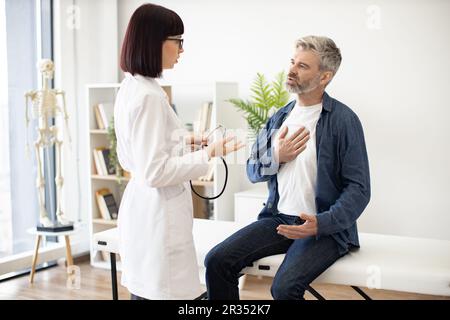 Besorgter, grauhaariger Mann, der auf der Untersuchungscouch sitzt, mit der Hand auf der Brust, während eine aufmerksame Frau im Mantel des Arztes ein Stethoskop hält. Männlicher Patient, der dem Facharzt in der Sprechstunde Symptome beschreibt. Stockfoto