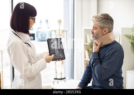 Eine weiße Dame hält ein Tablet mit CT-Scan-Bild auf dem Bildschirm, während sie mit einem Mann in Nackenstütze spricht, der auf dem Untersuchungstisch sitzt. Junger Orthopäde zur Analyse der Brustwirbelsäule auf einem digitalen Bericht in der Klinik. Stockfoto