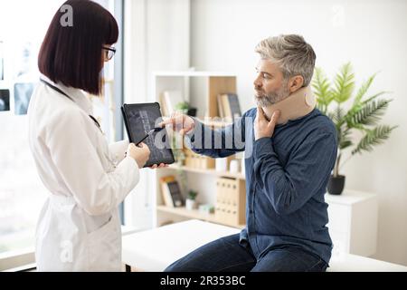 Eine weiße Dame hält ein Tablet mit CT-Scan-Bild auf dem Bildschirm, während sie mit einem Mann in Nackenstütze spricht, der auf dem Untersuchungstisch sitzt. Junger Orthopäde zur Analyse der Brustwirbelsäule auf einem digitalen Bericht in der Klinik. Stockfoto