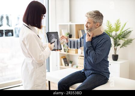 Eine weiße Dame hält ein Tablet mit CT-Scan-Bild auf dem Bildschirm, während sie mit einem Mann in Nackenstütze spricht, der auf dem Untersuchungstisch sitzt. Junger Orthopäde zur Analyse der Brustwirbelsäule auf einem digitalen Bericht in der Klinik. Stockfoto