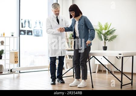 Selbstbewusster Mann im Labormantel, der einer schönen Frau in der Halskrause hilft, beim Gehen mit dem Stock in der Klinik das Gleichgewicht zu finden. Physiotherapeut unterstützt die Dame bei ihren ersten Schritten nach der Verletzung. Stockfoto