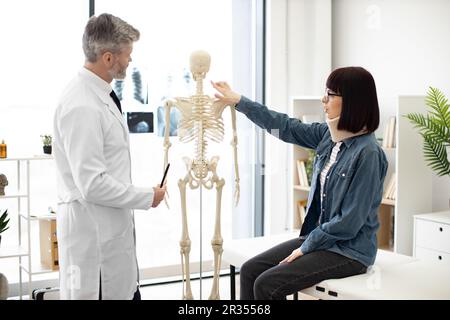 Seitenansicht eines erwachsenen Mannes im Arztmantel, der auf das Skelett zeigt, während eine junge Frau mit Brille und Halsband auf der Couch sitzt. Hausarzt zur Darstellung von Halsverletzungen am anatomischen Modell in der Klinik. Stockfoto