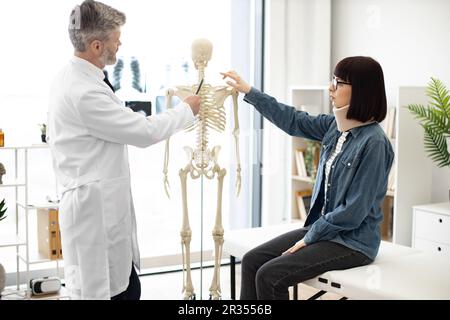Seitenansicht eines erwachsenen Mannes im Arztmantel, der auf das Skelett zeigt, während eine junge Frau mit Brille und Halsband auf der Couch sitzt. Hausarzt zur Darstellung von Halsverletzungen am anatomischen Modell in der Klinik. Stockfoto