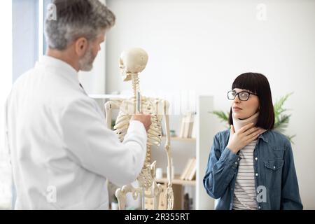 Schulterblick einer schönen jungen Frau mit Halsspange und Blick auf das menschliche Skelett eines erwachsenen Mannes. Beunruhigte Frau in Brille, die nach einer Wirbelsäulenverletzung einen Arzt zur Beratung besucht. Stockfoto