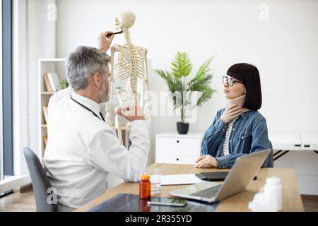 Eine charmante Frau, die am Schreibtisch des Arztes sitzt, während ein selbstbewusster Mann im weißen Mantel auf die Halswirbelsäule des Skeletts zeigt. Junge Frau mit Halsverletzung beim Hausarzt durch Termin in der Klinik. Stockfoto