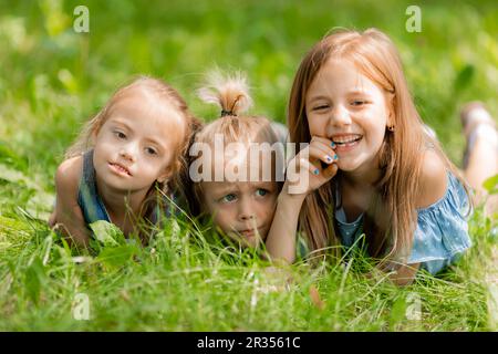 Drei kleine Kinder liegen im Sommer im grünen Gras. Glückliche Kindheit. Ein Kind mit Down-Syndrom. Hochwertiges Foto Stockfoto