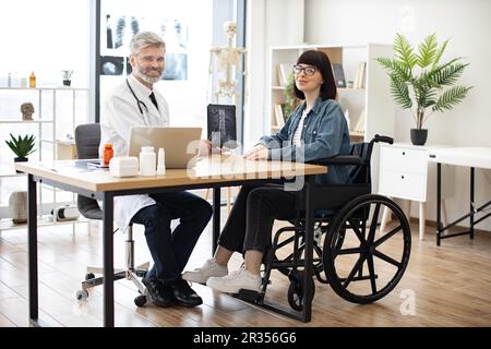 Volllängenporträt einer freundlichen Ärztin und einer glücklichen behinderten Frau bei einem Gespräch im Sprechzimmer der Klinik. Fröhlicher Arzt zeigt Röntgenaufnahmen auf Tablet mit positiven Ergebnissen bei einer Frau im Rollstuhl. Stockfoto