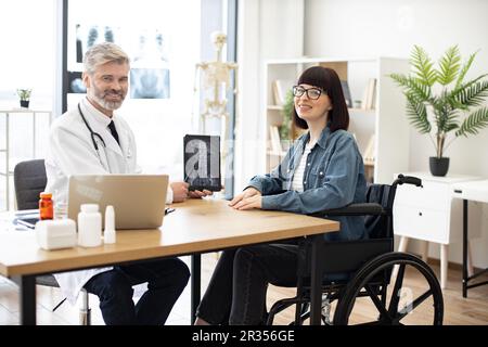 Volllängenporträt einer freundlichen Ärztin und einer glücklichen behinderten Frau bei einem Gespräch im Sprechzimmer der Klinik. Fröhlicher Arzt zeigt Röntgenaufnahmen auf Tablet mit positiven Ergebnissen bei einer Frau im Rollstuhl. Stockfoto