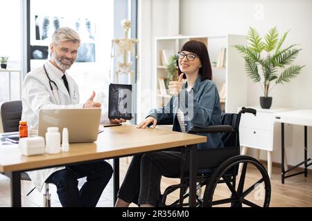 Volllängenporträt einer freundlichen Ärztin und einer glücklichen behinderten Frau bei einem Gespräch im Sprechzimmer der Klinik. Fröhlicher Arzt zeigt Röntgenaufnahmen auf Tablet mit positiven Ergebnissen bei einer Frau im Rollstuhl. Stockfoto